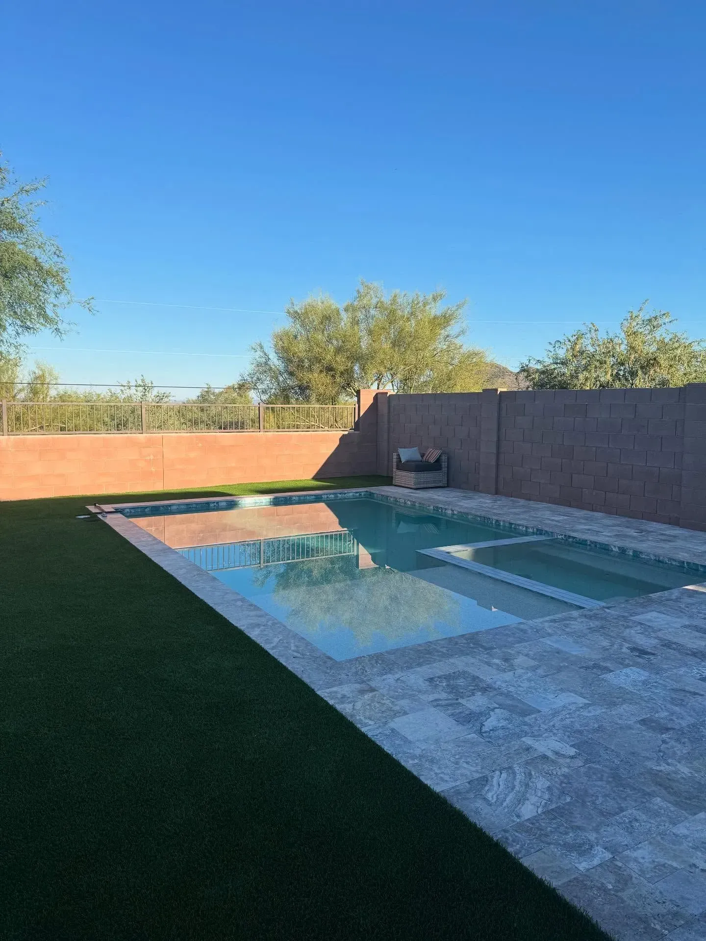 Rectangular pool with clear water, surrounded by green turf and stone patio. Brown brick wall and clear sky in background.