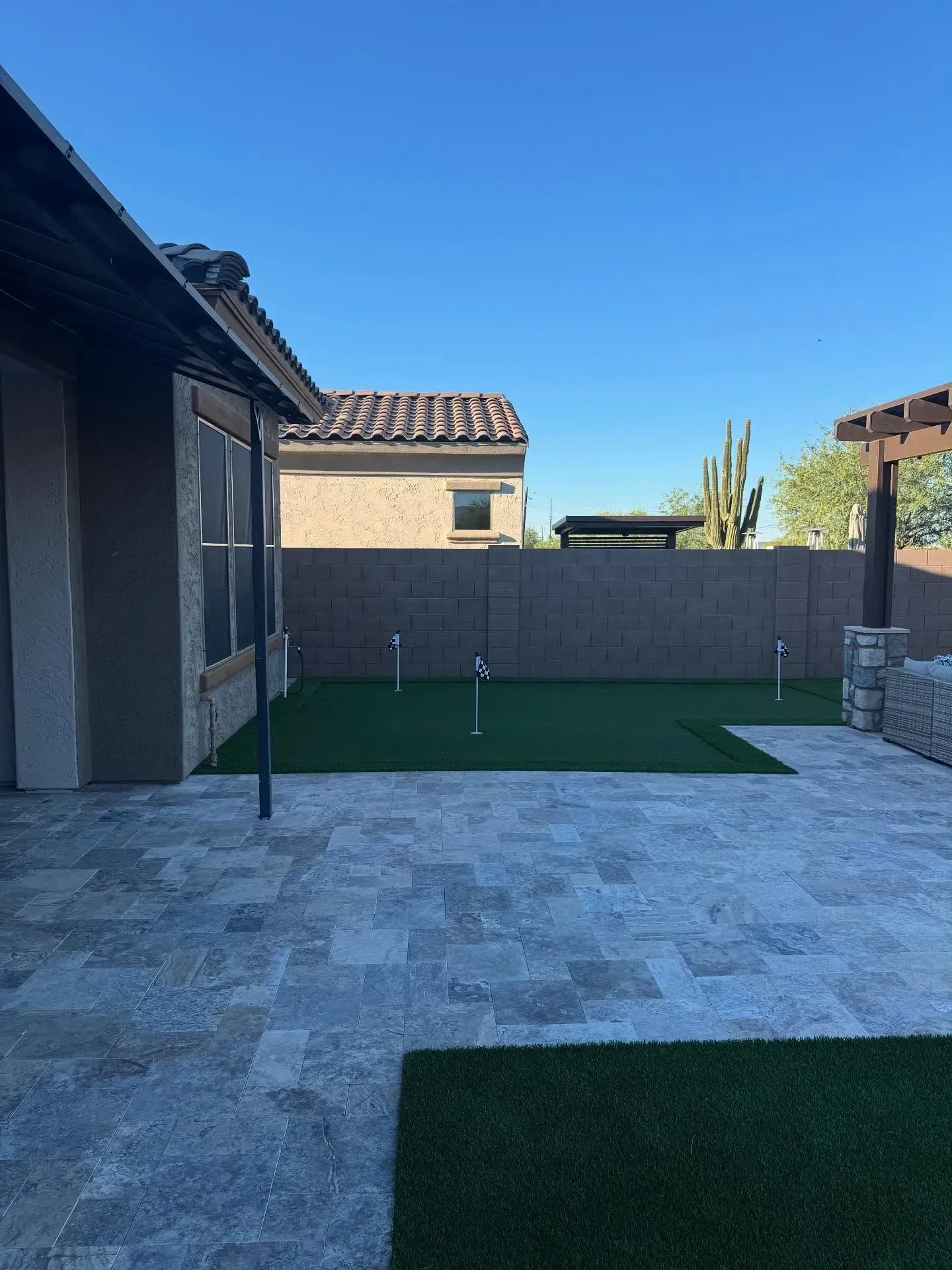 Backyard patio with gray stone pavers, green turf, and a brown stucco house under a blue sky.