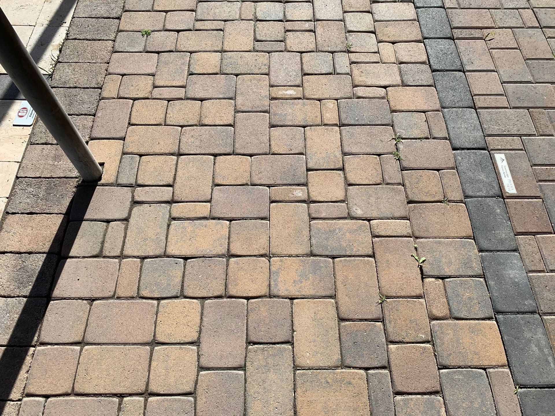 Brick pavers on a sidewalk in varying shades of brown and gray. A metal pole casts a shadow.