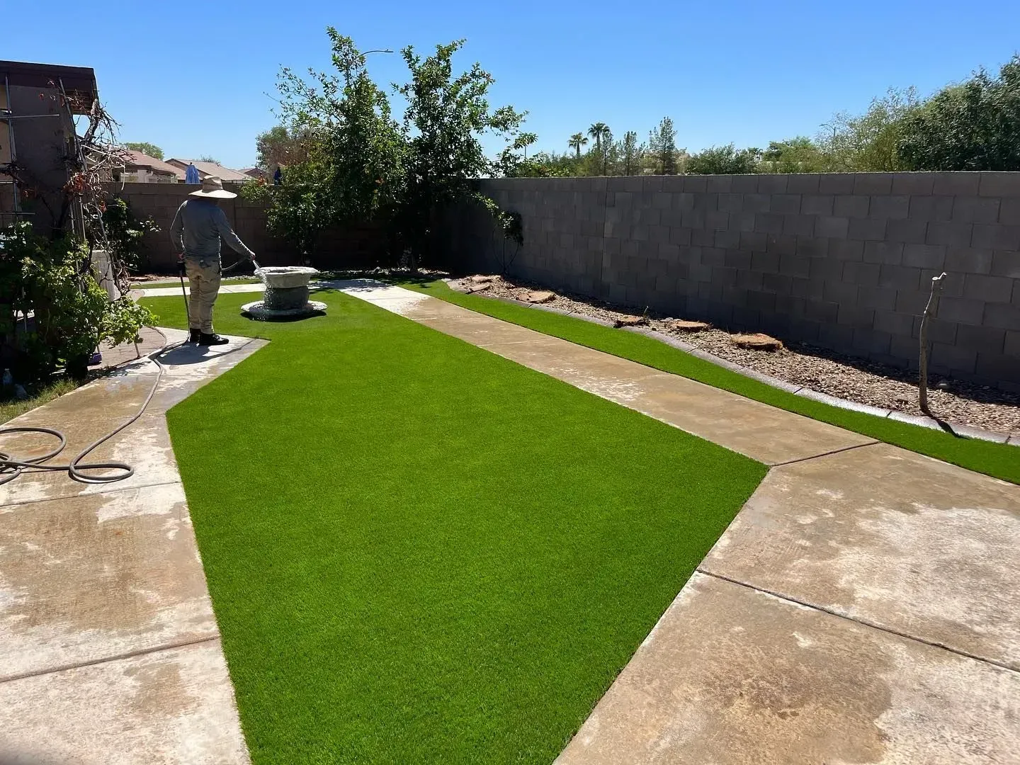 A person sprays water on artificial turf in a backyard with concrete paths and a retaining wall.
