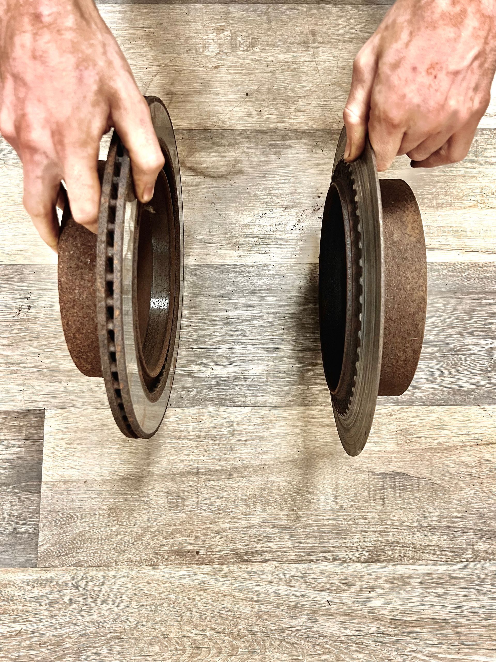 Hands holding two weathered, circular car brake rotors against a wood-grain background.