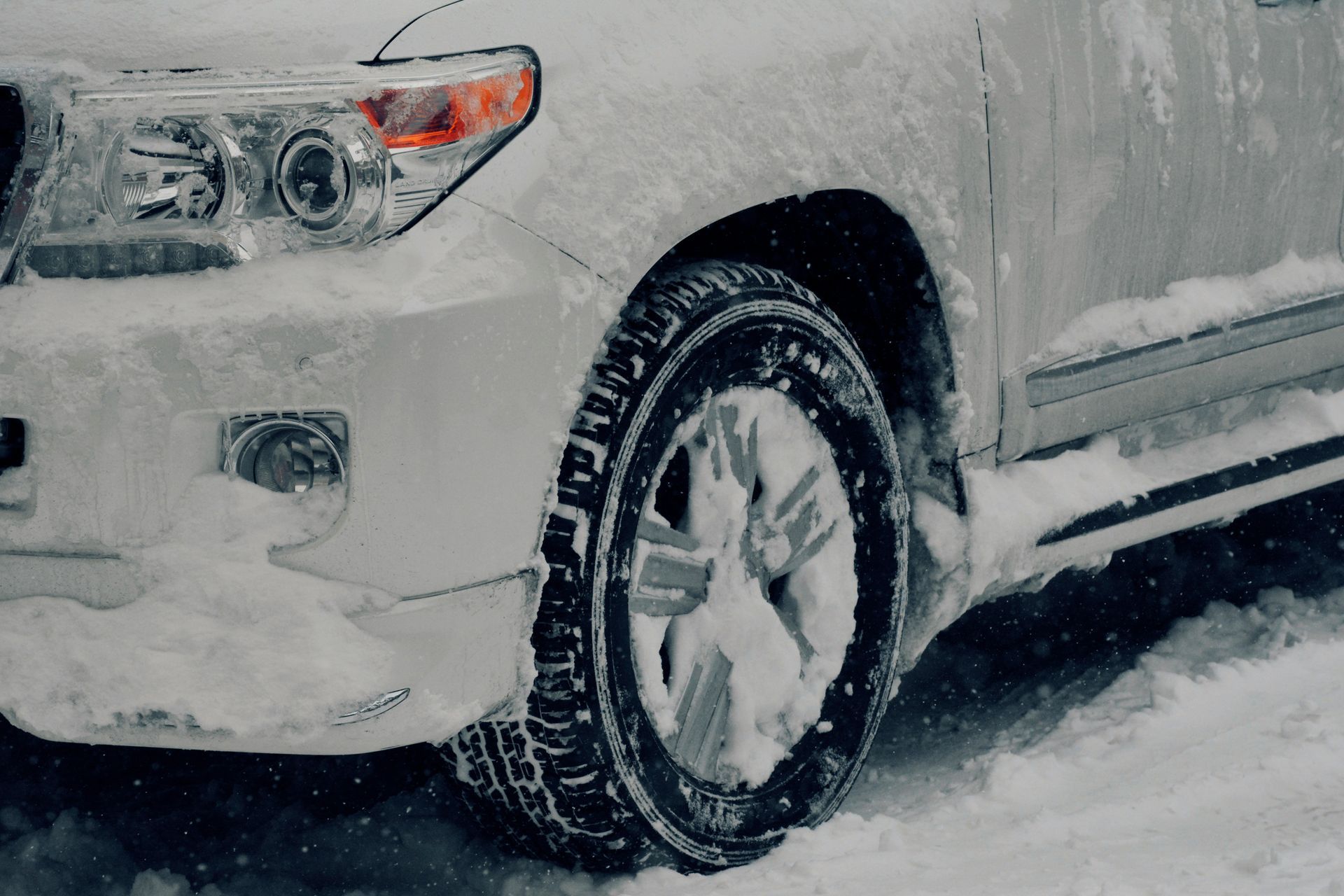 A close-up of a white SUV's front wheel and bumper covered in fresh snow during a winter storm.
