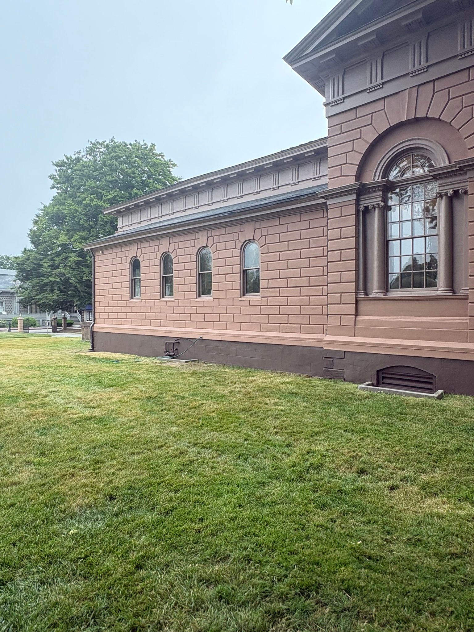 Side view of a historic brick building with arched windows, a grassy lawn, and cloudy sky.