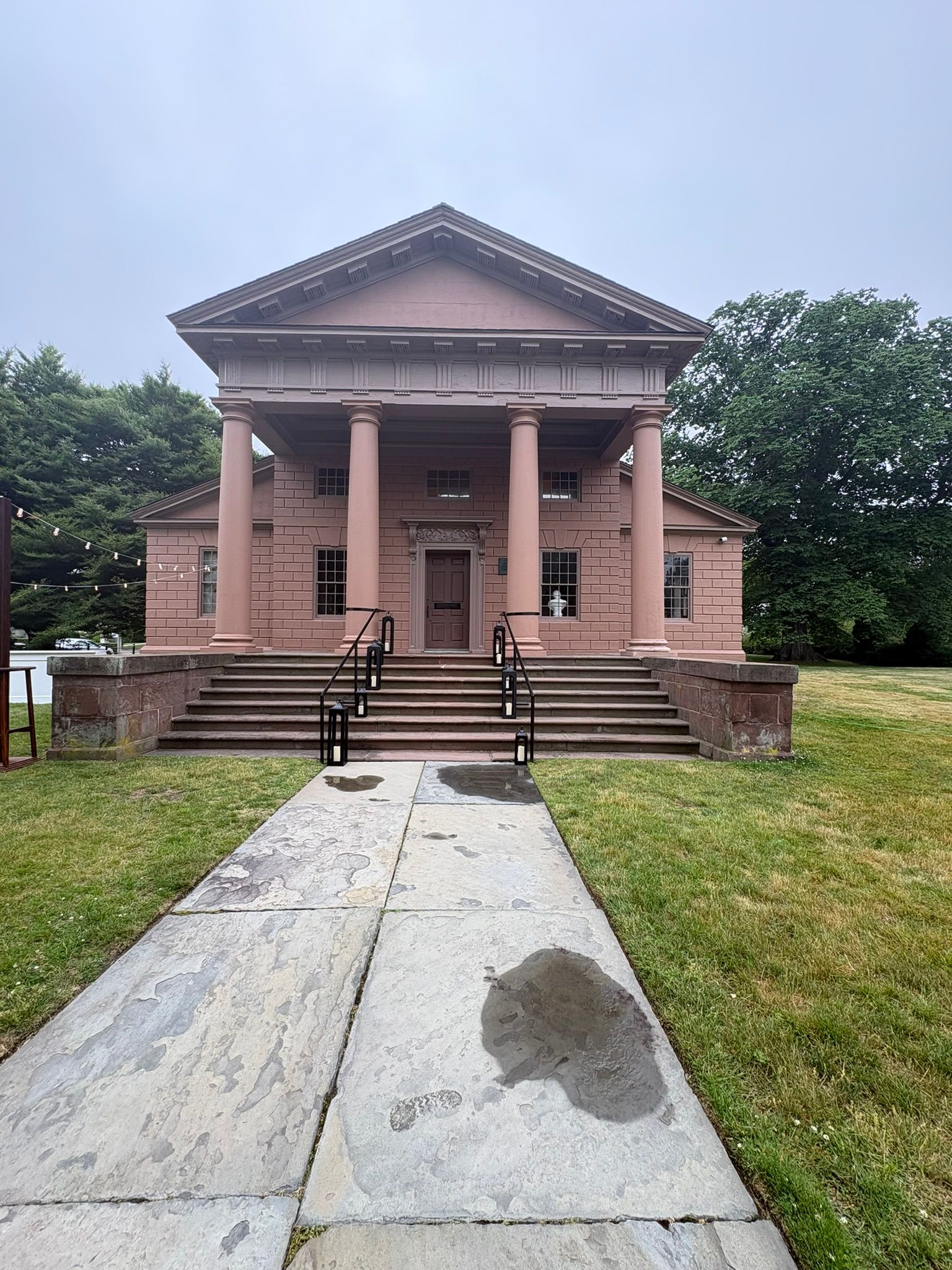 Brick building with columns and steps at the end of a wet concrete path, surrounded by grass and trees.