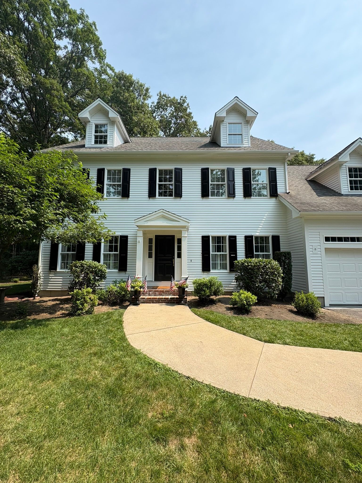 White two-story house with black shutters, dormer windows, a front porch, and a curved walkway under trees