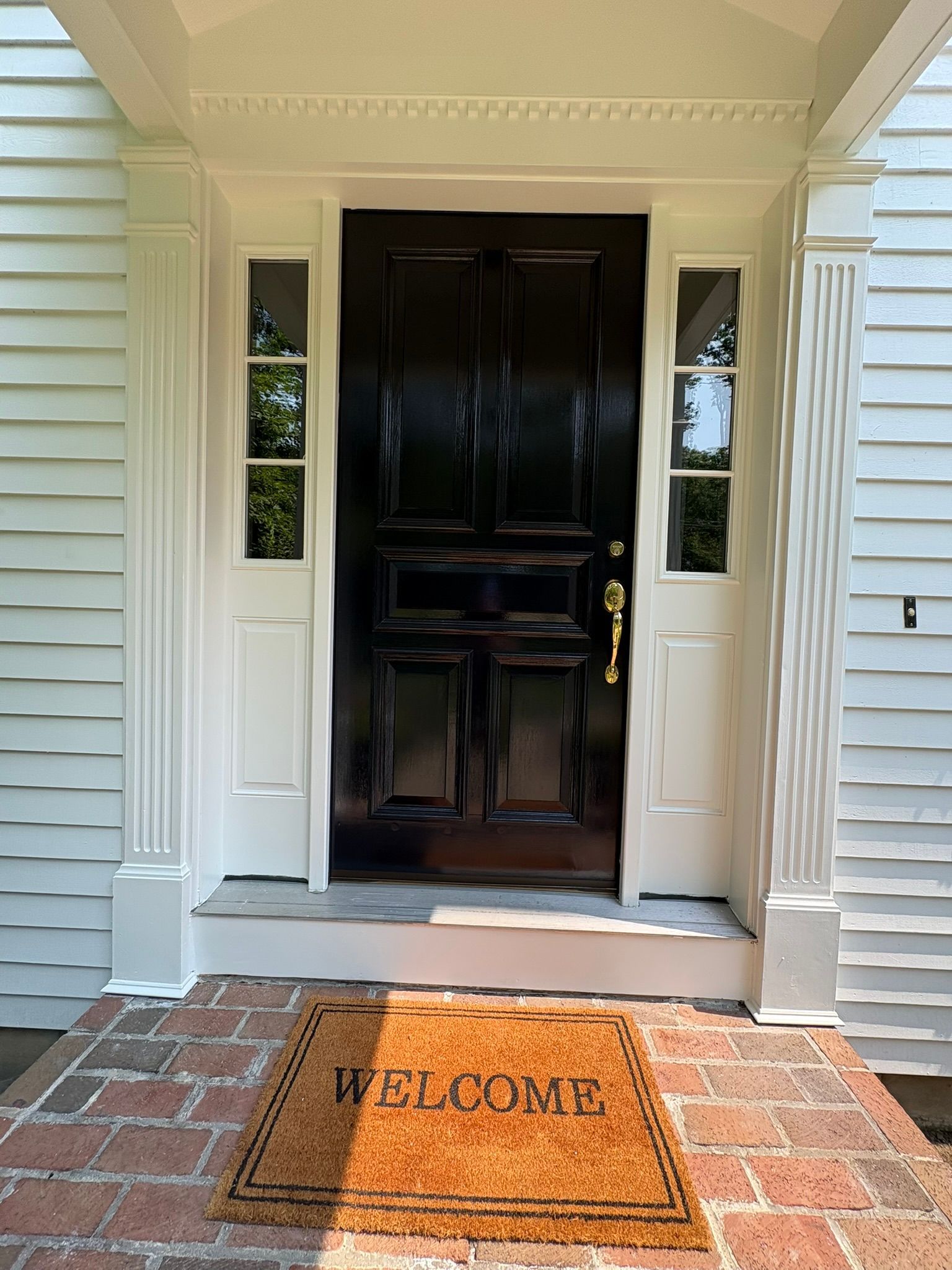 Front porch with a black front door, white columns, and a welcome mat on a brick entryway.