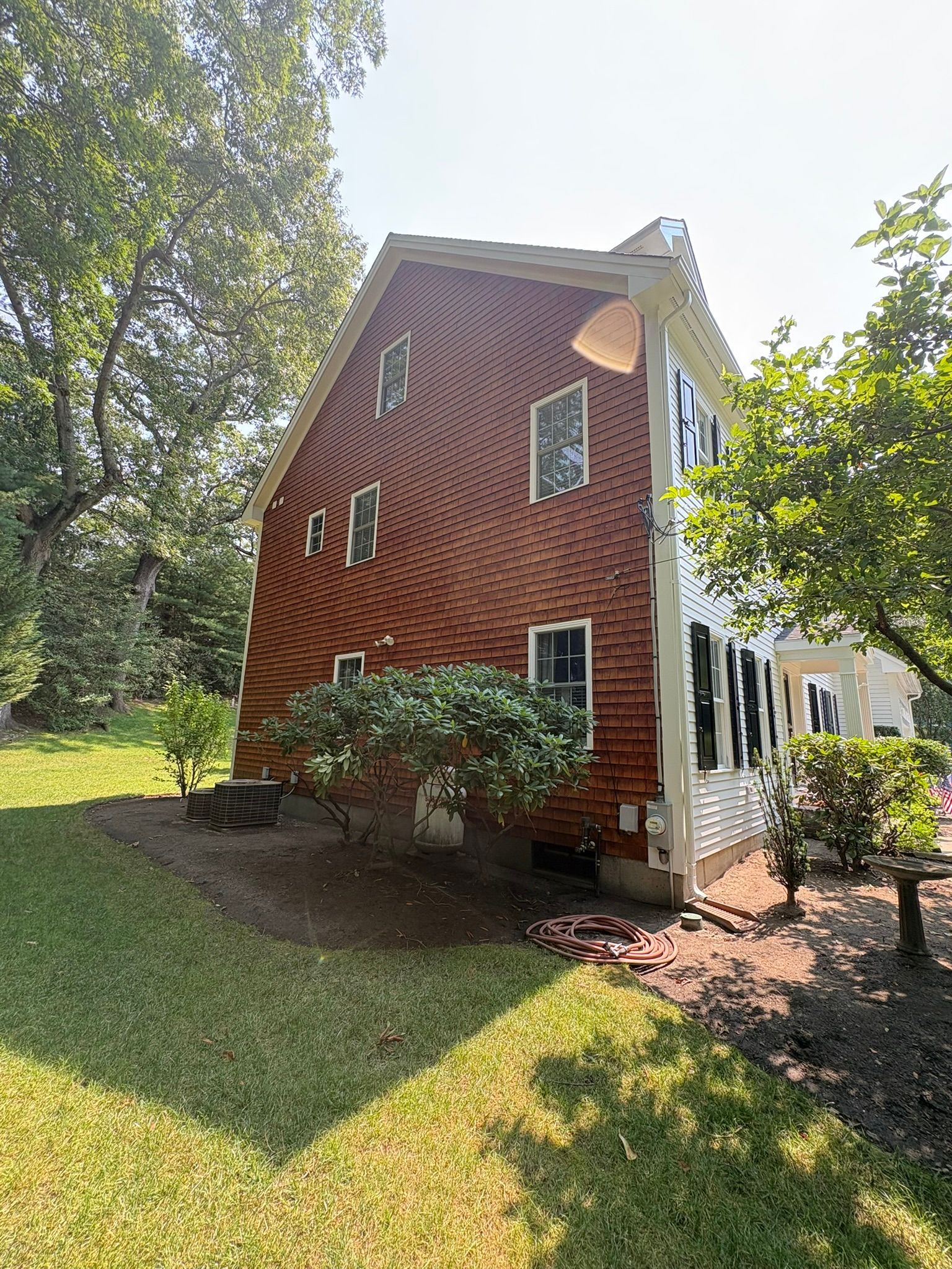 Red shingled house exterior with white trim, small windows, and trees casting shade on the lawn.