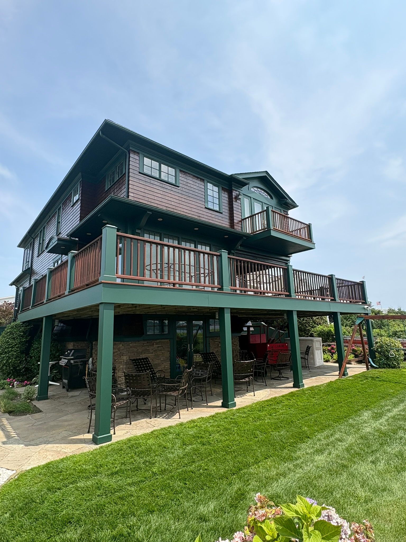 Two-story house on stilts with green trim and wraparound deck on a grassy hillside under a blue sky