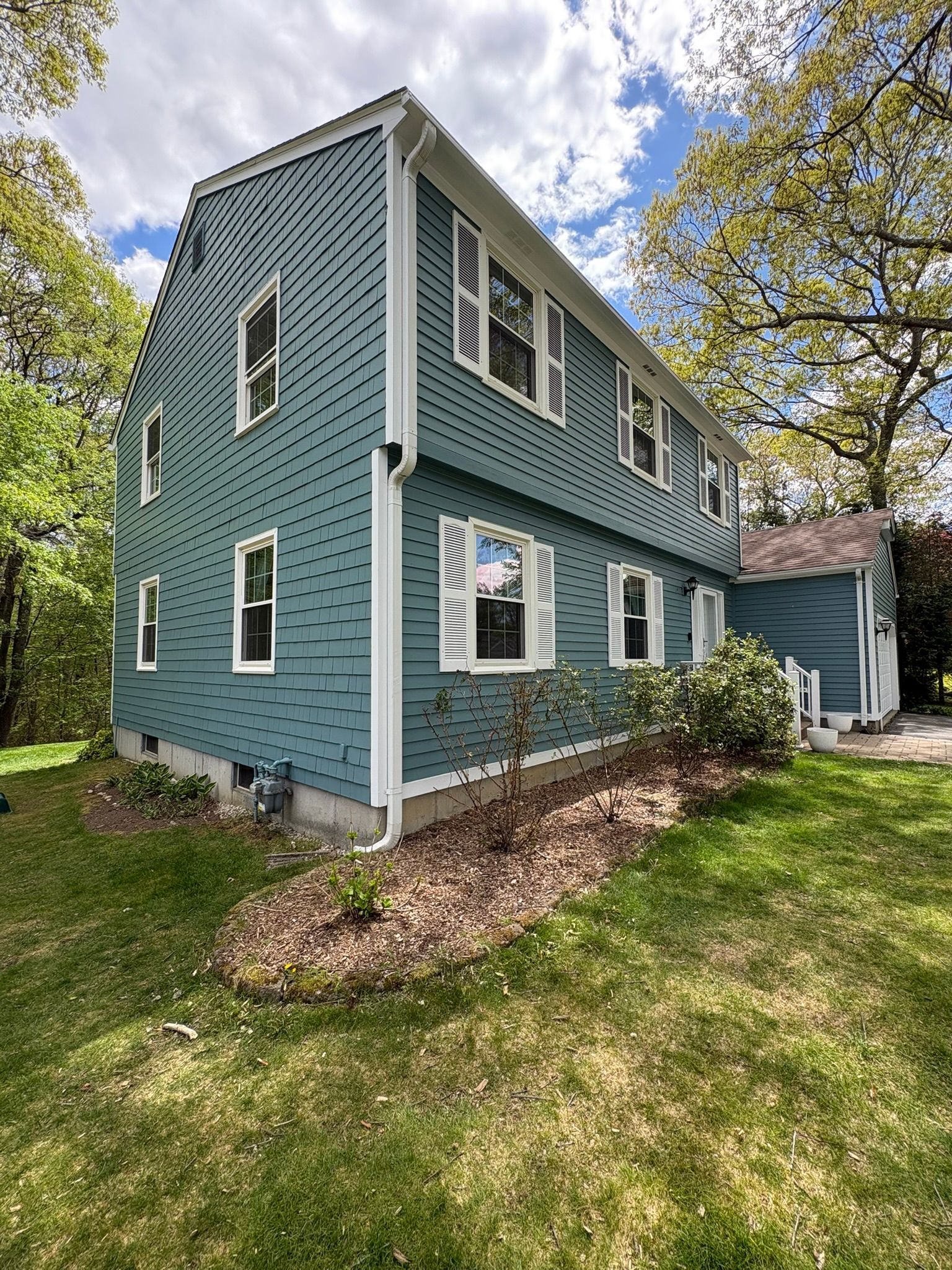 Blue two-story house with white trim, dormer windows, and a small side porch amid trees and grass.