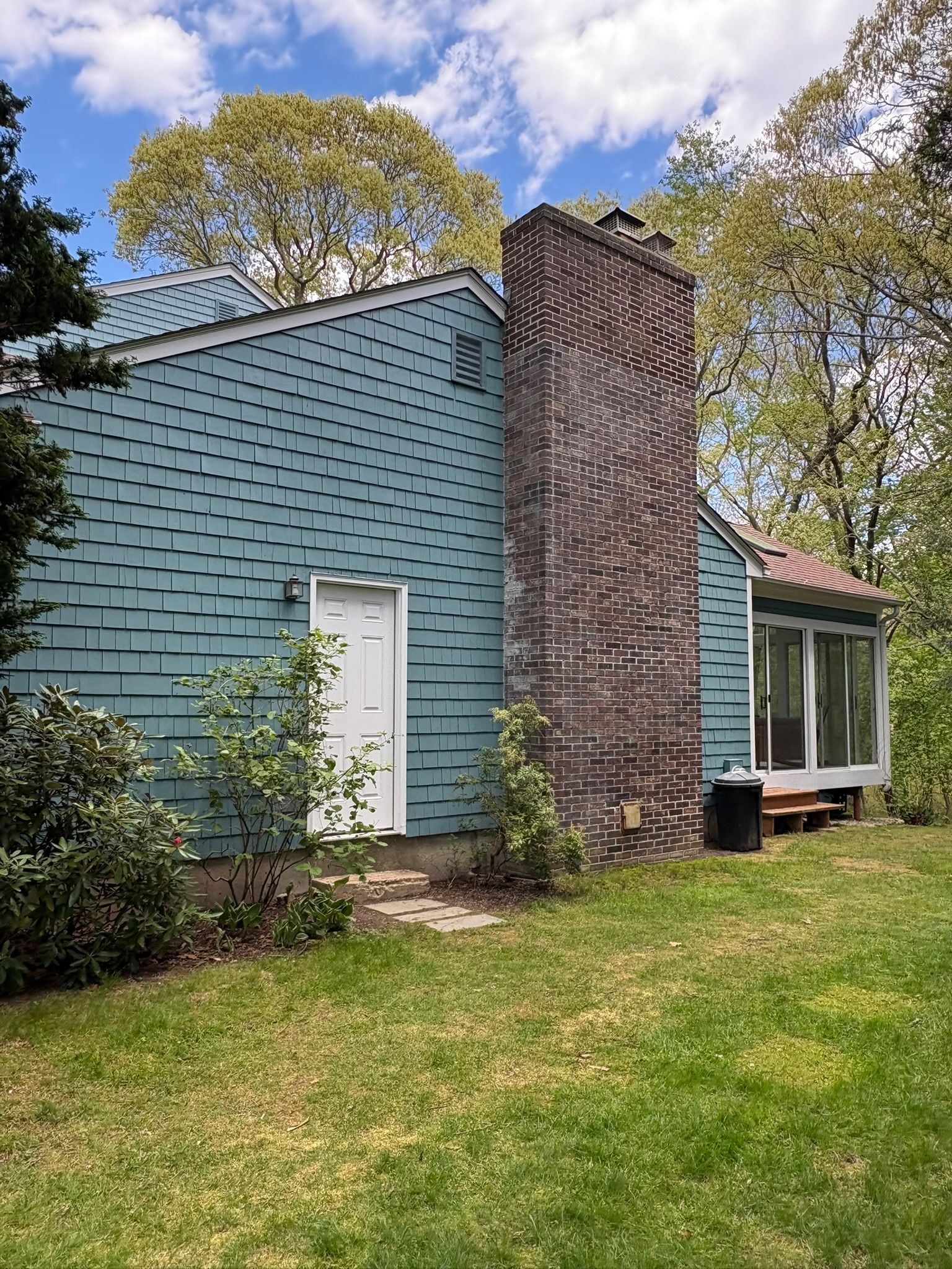 Blue shingled house with a brick chimney and white door, seen from a grassy yard under a partly cloudy sky