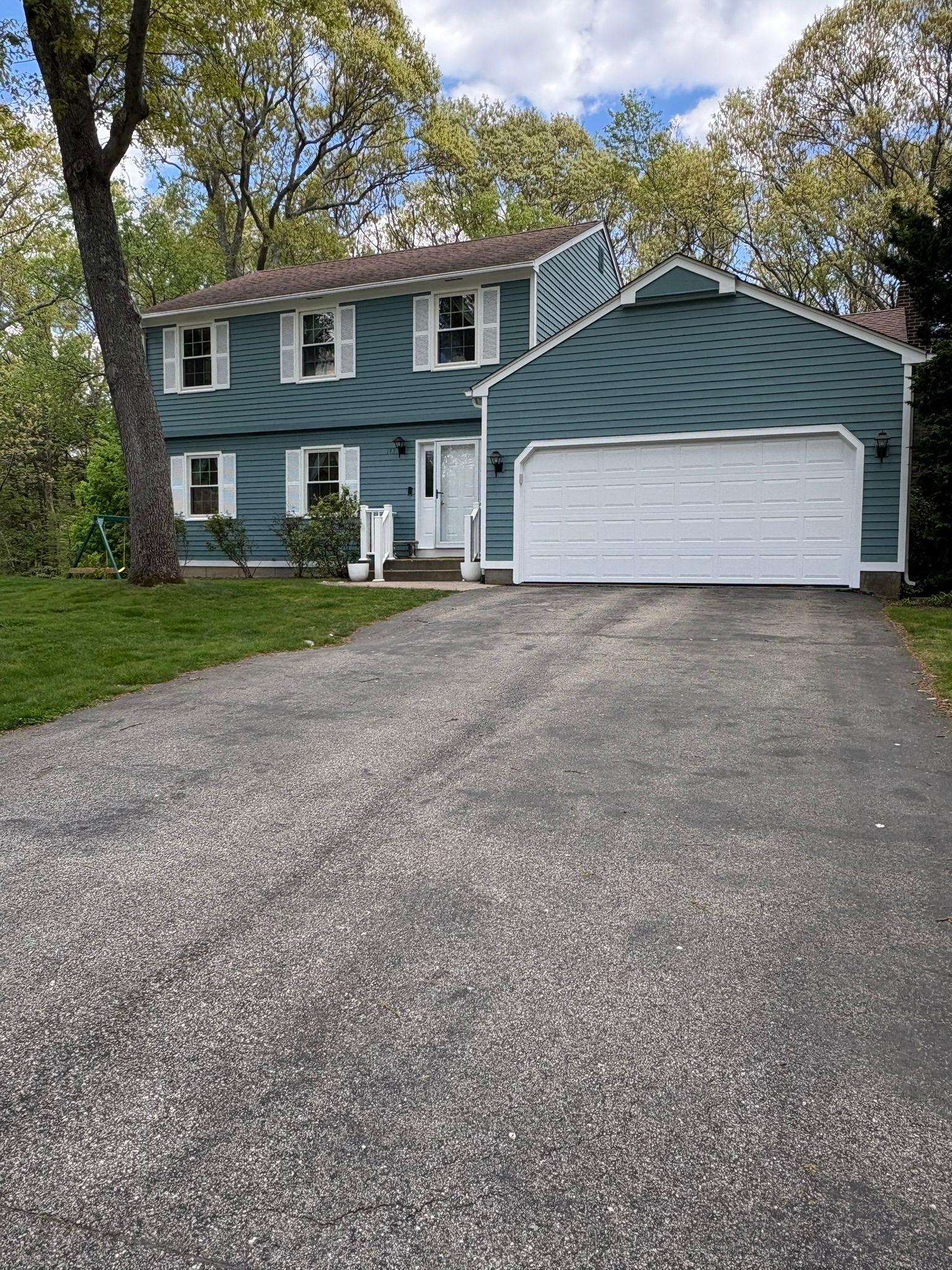 Blue two-story house with white garage and gravel driveway, surrounded by trees.