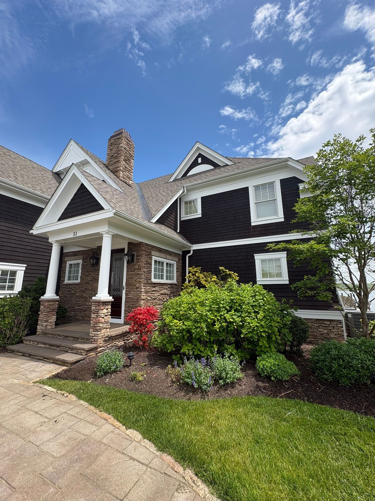 Brick and dark shingle house with white trim, front porch, stone chimney, and landscaped yard under a blue sky
