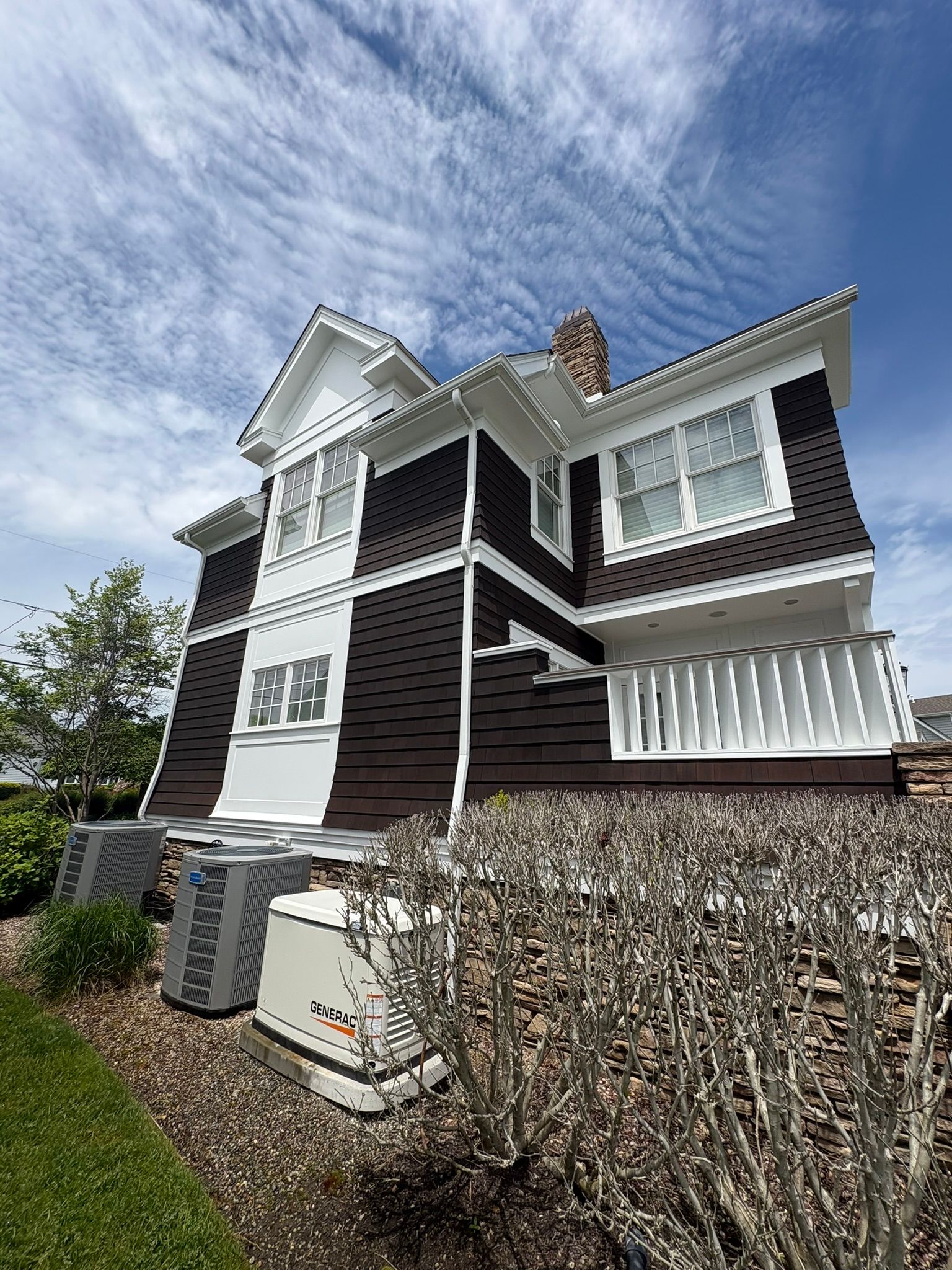 Two-story dark house with white trim and hedges under a cloudy blue sky