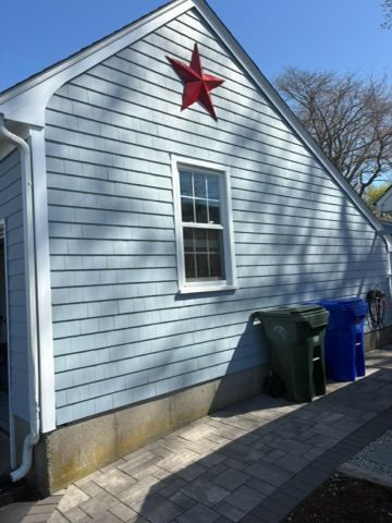 Light blue house side with a red star, window, trash bins, and paved walkway.