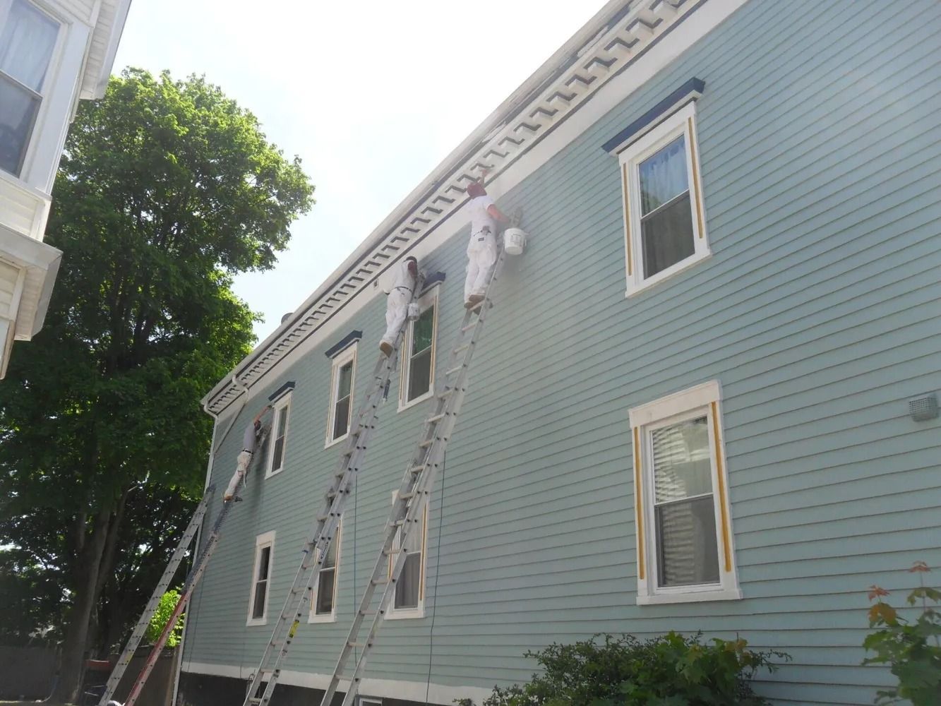 Two painters on ladders painting a light blue house trim. Sunny day, exterior shot.