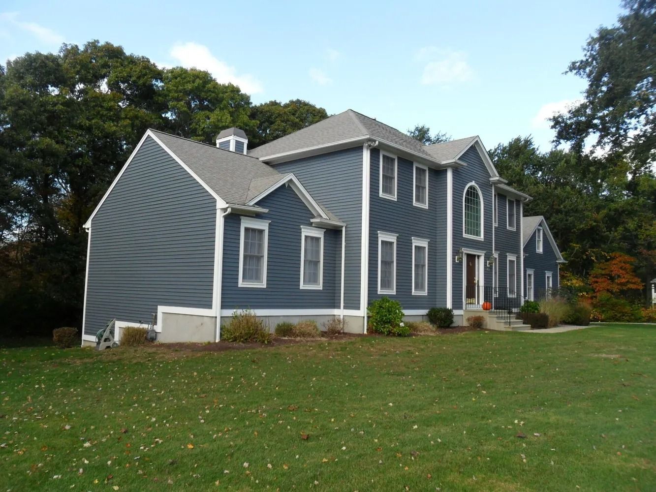 Blue house with white trim, green lawn, and surrounding trees under a blue sky.