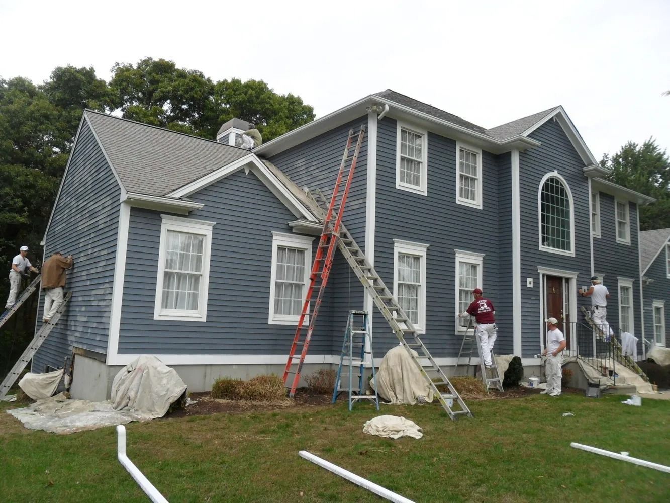 House being painted blue with white trim. Several workers on ladders, green yard.