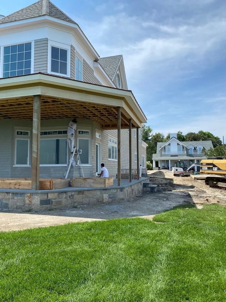 House under construction with workers, stone facade, covered porch, and blue sky.