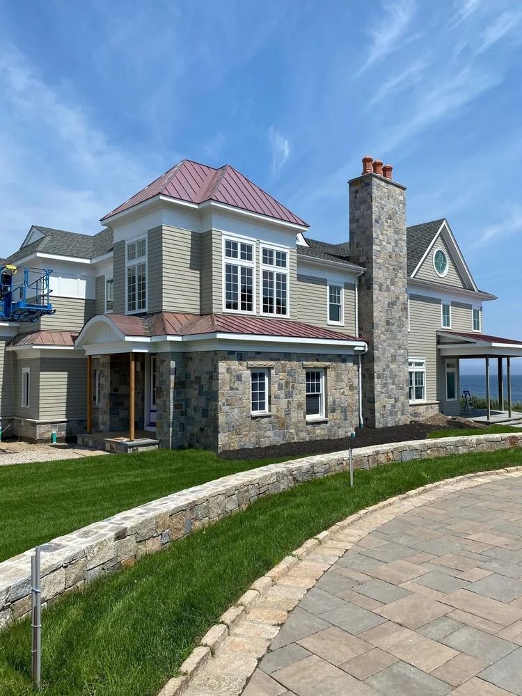 Two-story house with stone and beige siding, red roof, and chimney. Overlooks ocean on a sunny day.