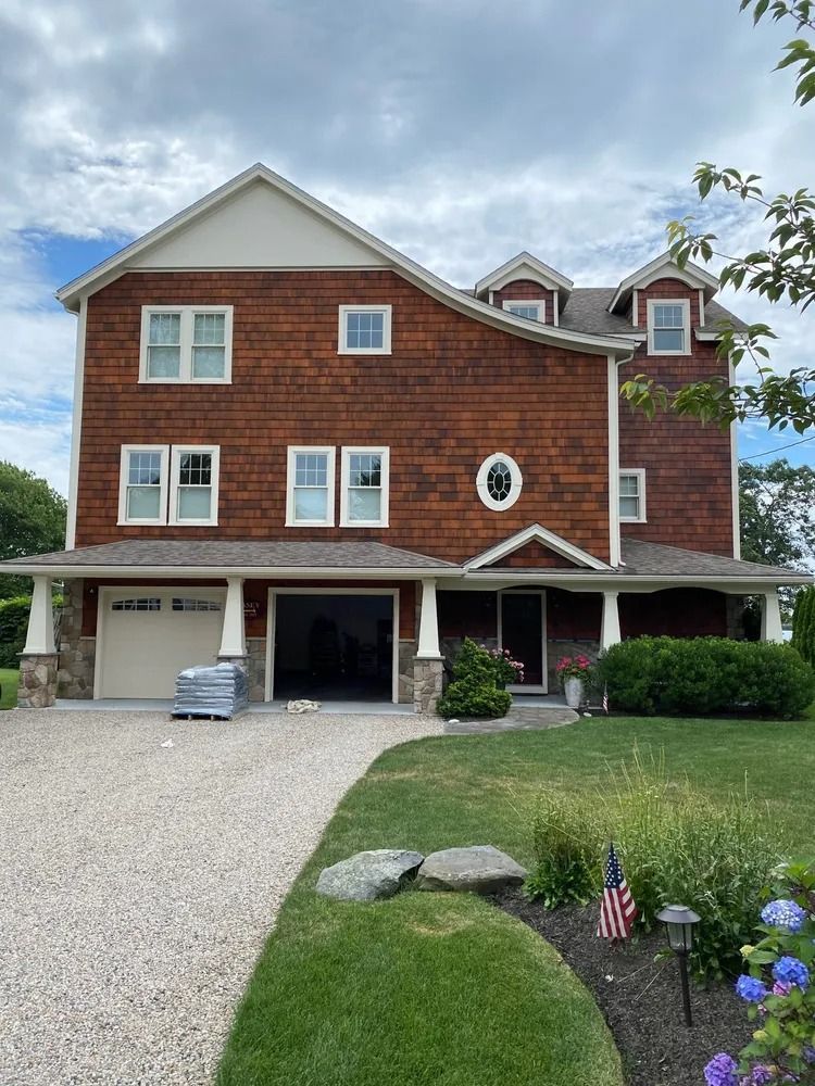 Brown shingled house with garage, porch, and gravel driveway under cloudy sky.
