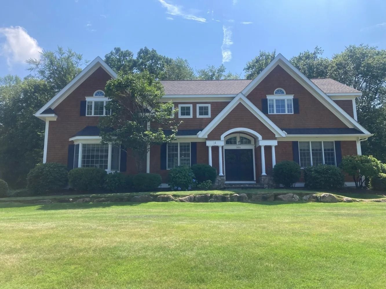 Two-story brick house with dark shutters, front door, and manicured lawn. Blue sky, surrounded by trees.