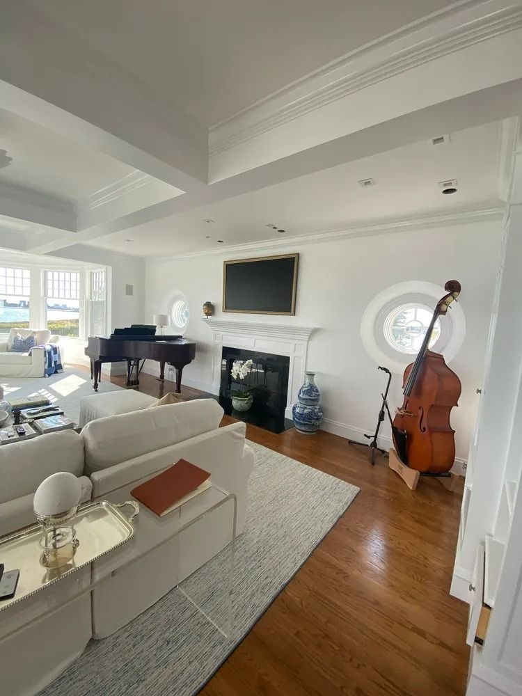 Living room with piano, fireplace, double bass, and large windows. White walls and ceiling, hardwood floors.