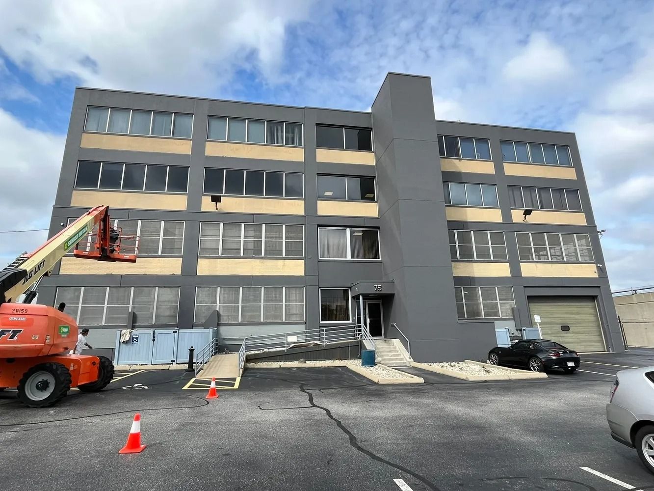 Gray industrial building with ramp, lift, and parked cars under a partly cloudy sky.