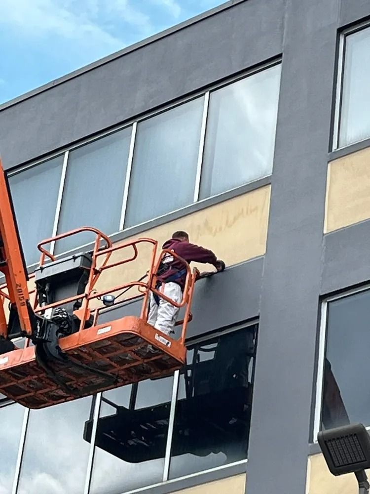Person in an orange lift working on a building exterior, which has dark gray trim and large windows.