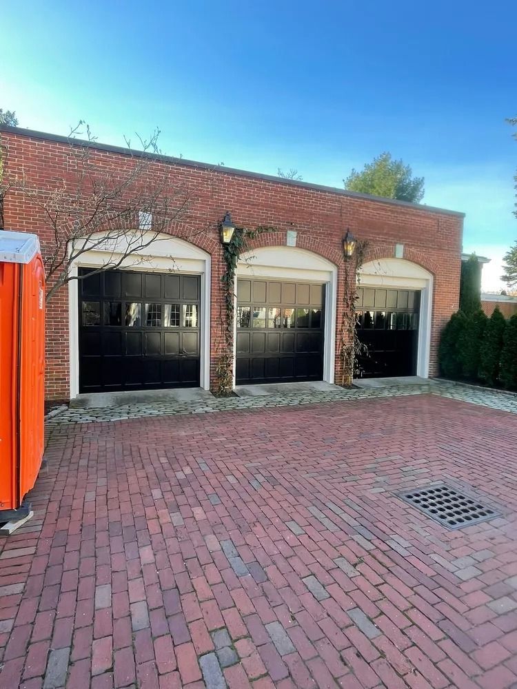 Brick building with three black garage doors, brick paved driveway, and orange portable toilet on the left.