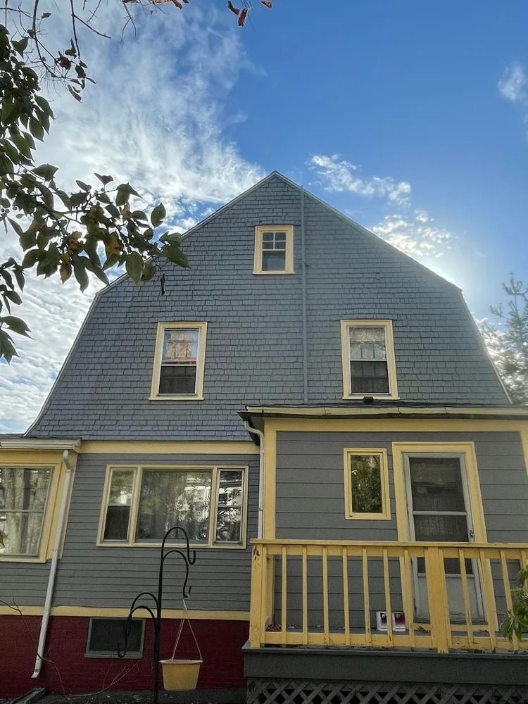 Gray house with a distinctive roof and yellow trim under a cloudy blue sky.