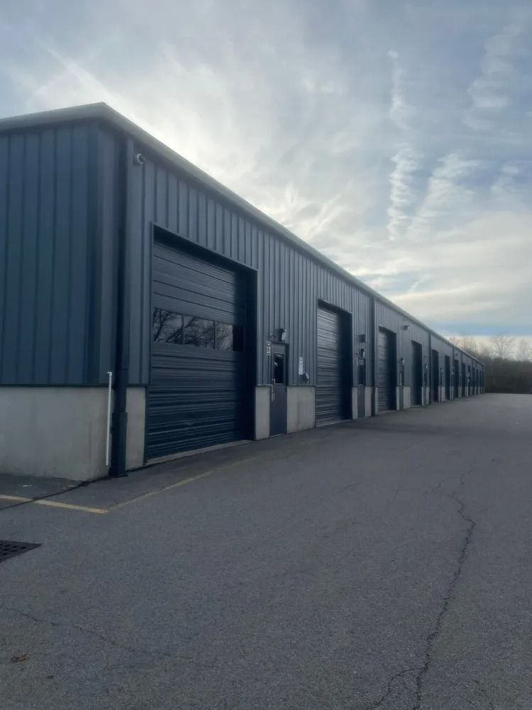 A row of dark blue industrial storage units with roll-up doors, on a gray asphalt lot under a cloudy sky.