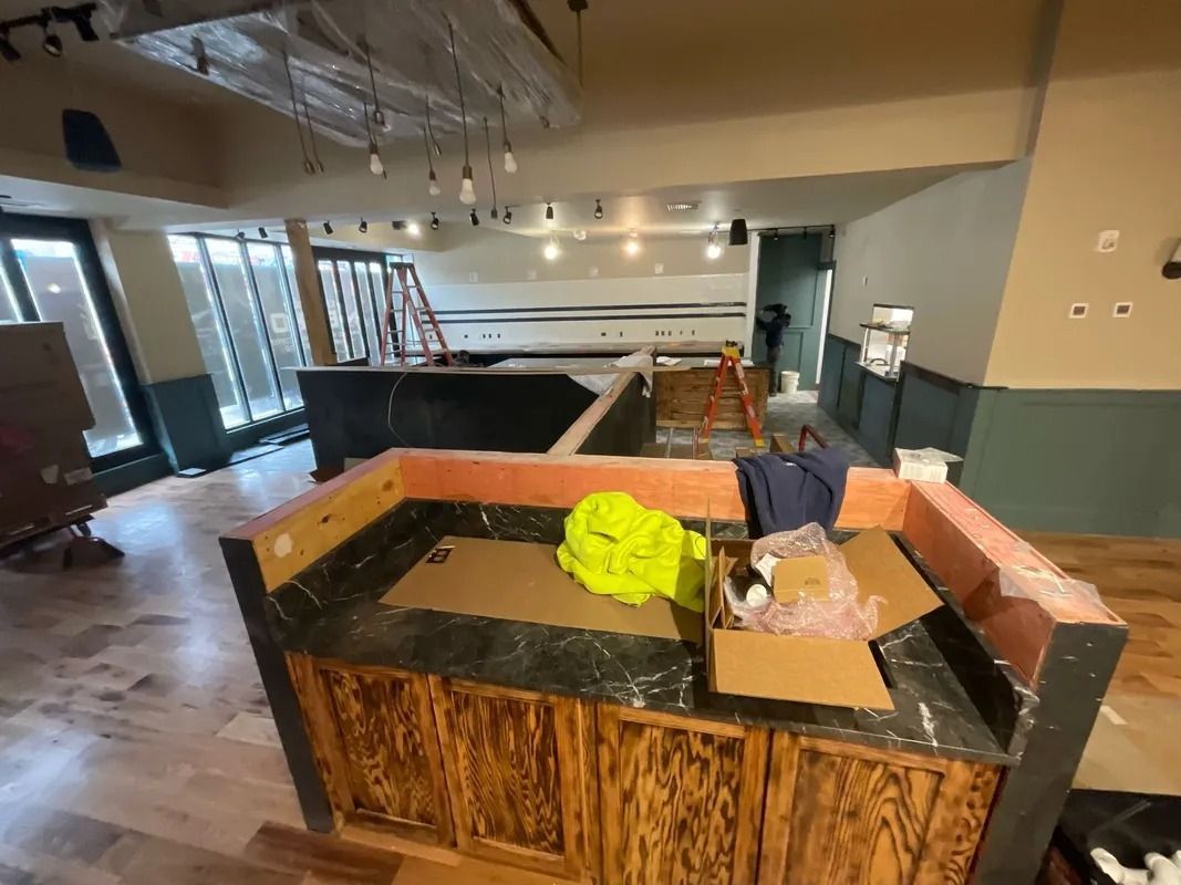 Interior of a restaurant under construction with a bar, wooden floors, and walls. Cardboard and construction materials are visible.