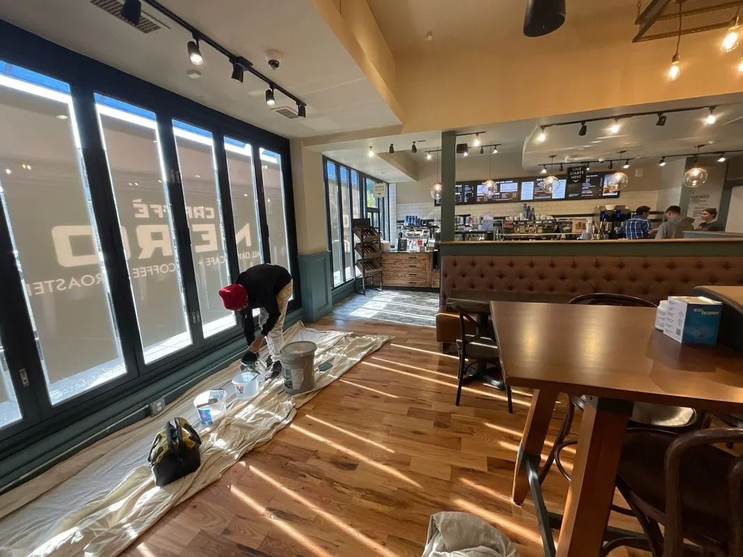 Person painting a coffee shop floor. Sunlight streams in. Baskets and supplies are present.
