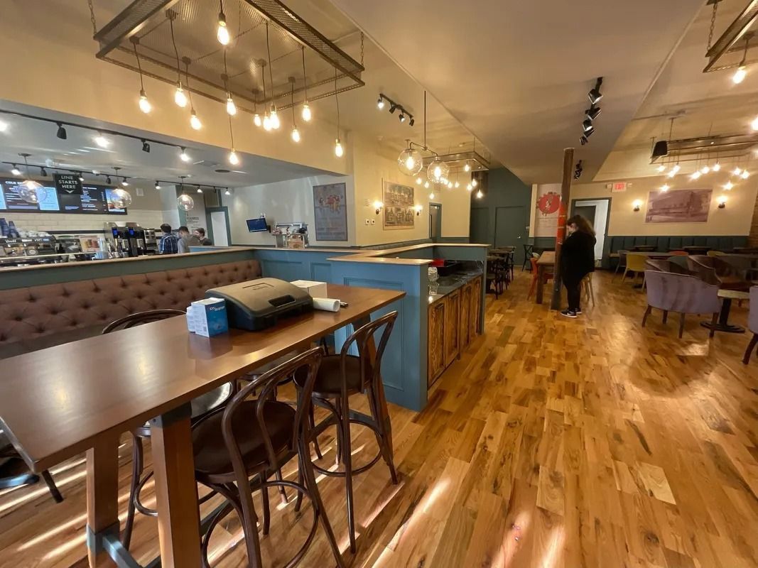 Restaurant interior with wooden floors, tables, and bar area. Overhead lighting and a person in the distance.