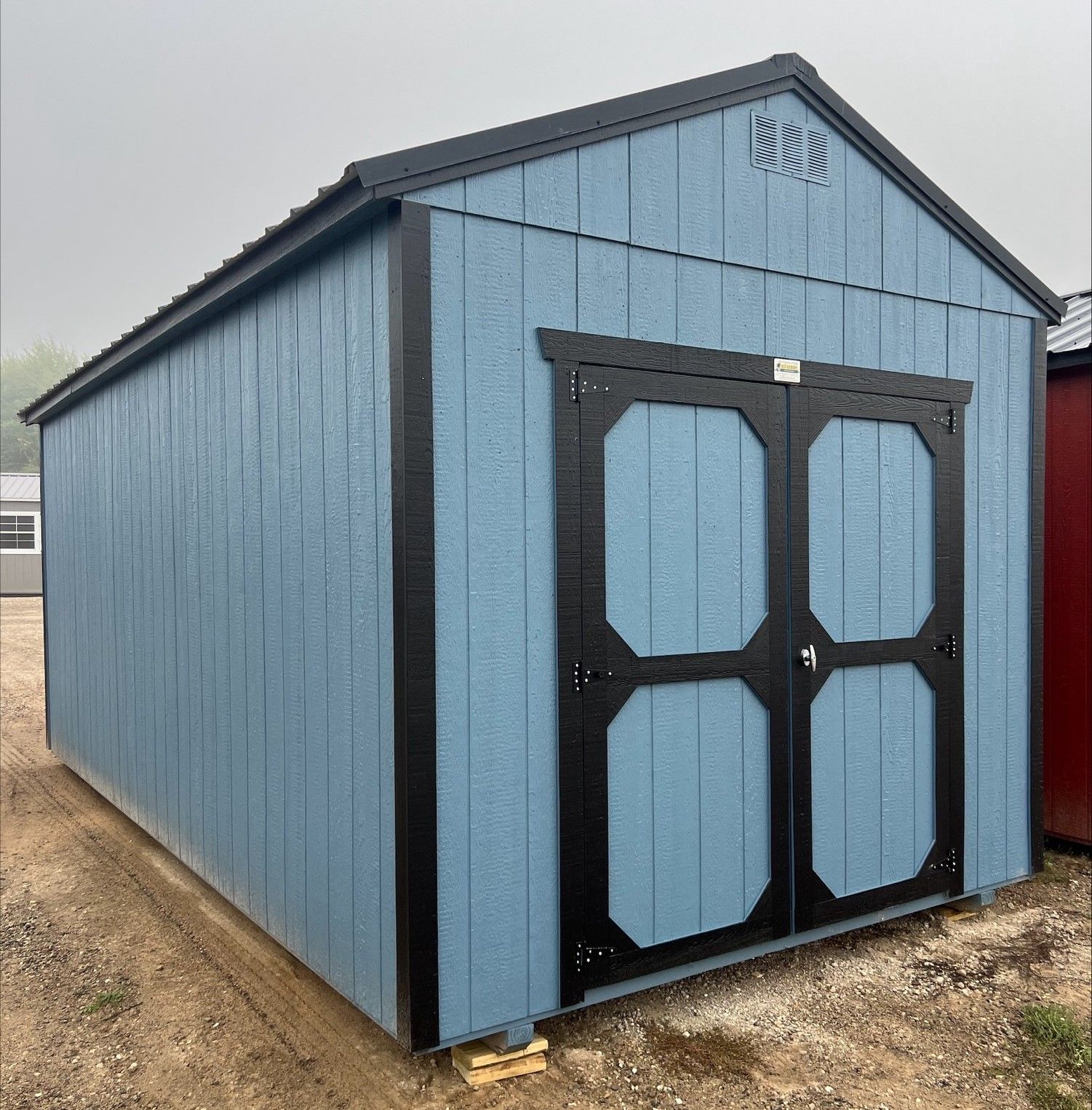 A red wooden storage shed with cream trim, a single window, and double doors, standing on a grassy outdoor lot.