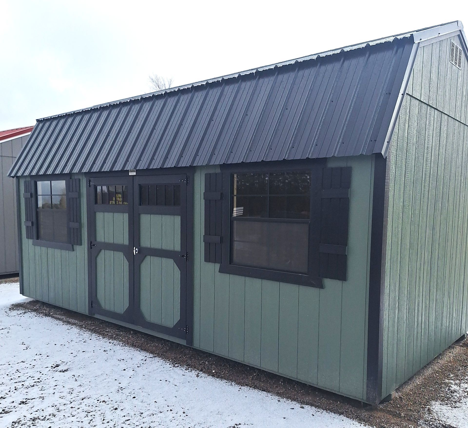 A grey, gabled garden shed with a white roller door and a side entry door, set on a gravel surface against a blue sky.