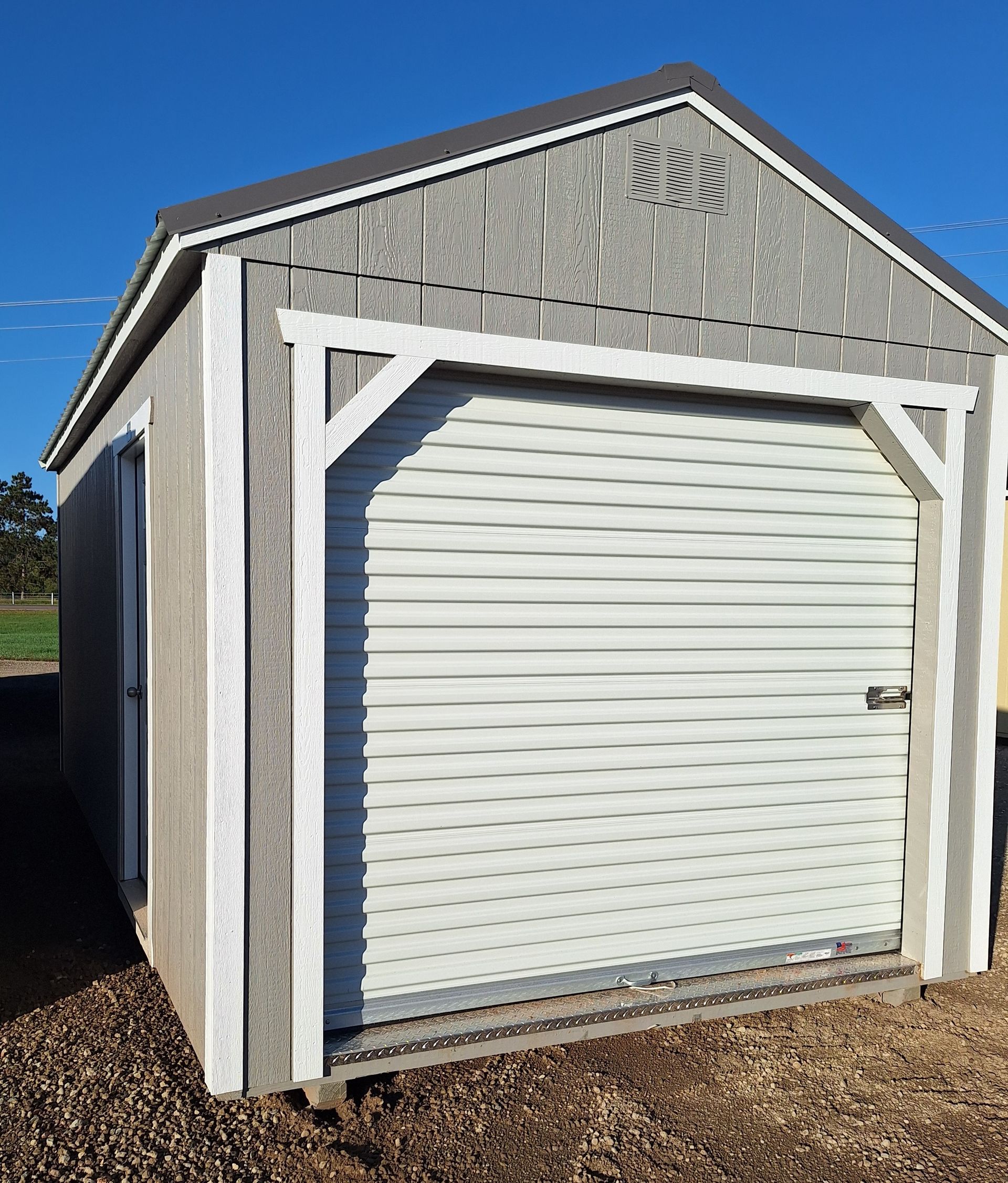 A grey, gabled garden shed with a white roller door and a side entry door, set on a gravel surface against a blue sky.