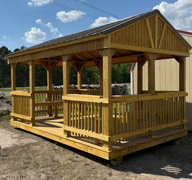 A wooden outdoor pavilion with a gabled roof, railings, and built-in benches, set on a dirt lot under a blue sky.