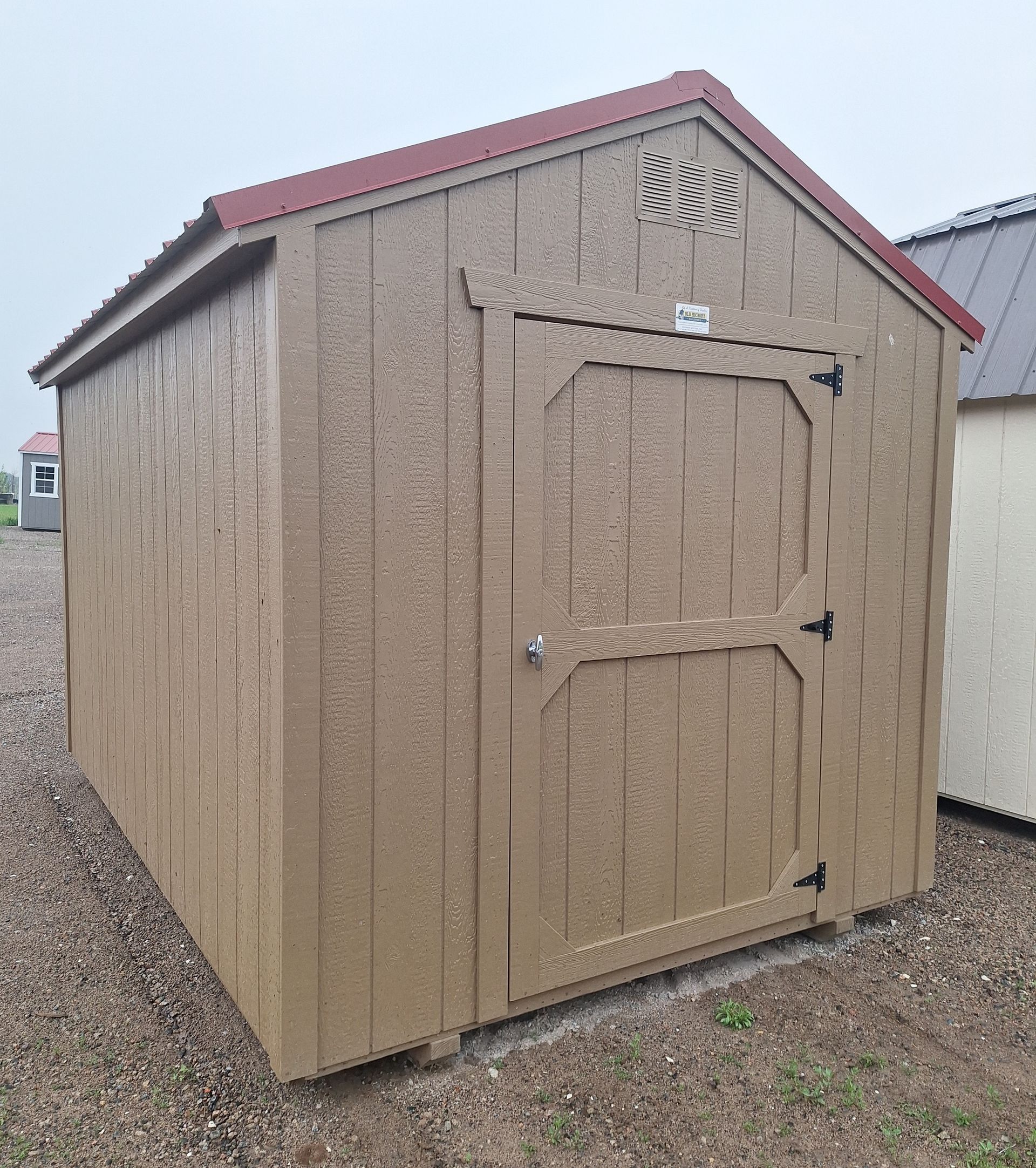 A tan wooden storage shed with a red metal roof and single door stands on a gravel lot under an overcast sky.