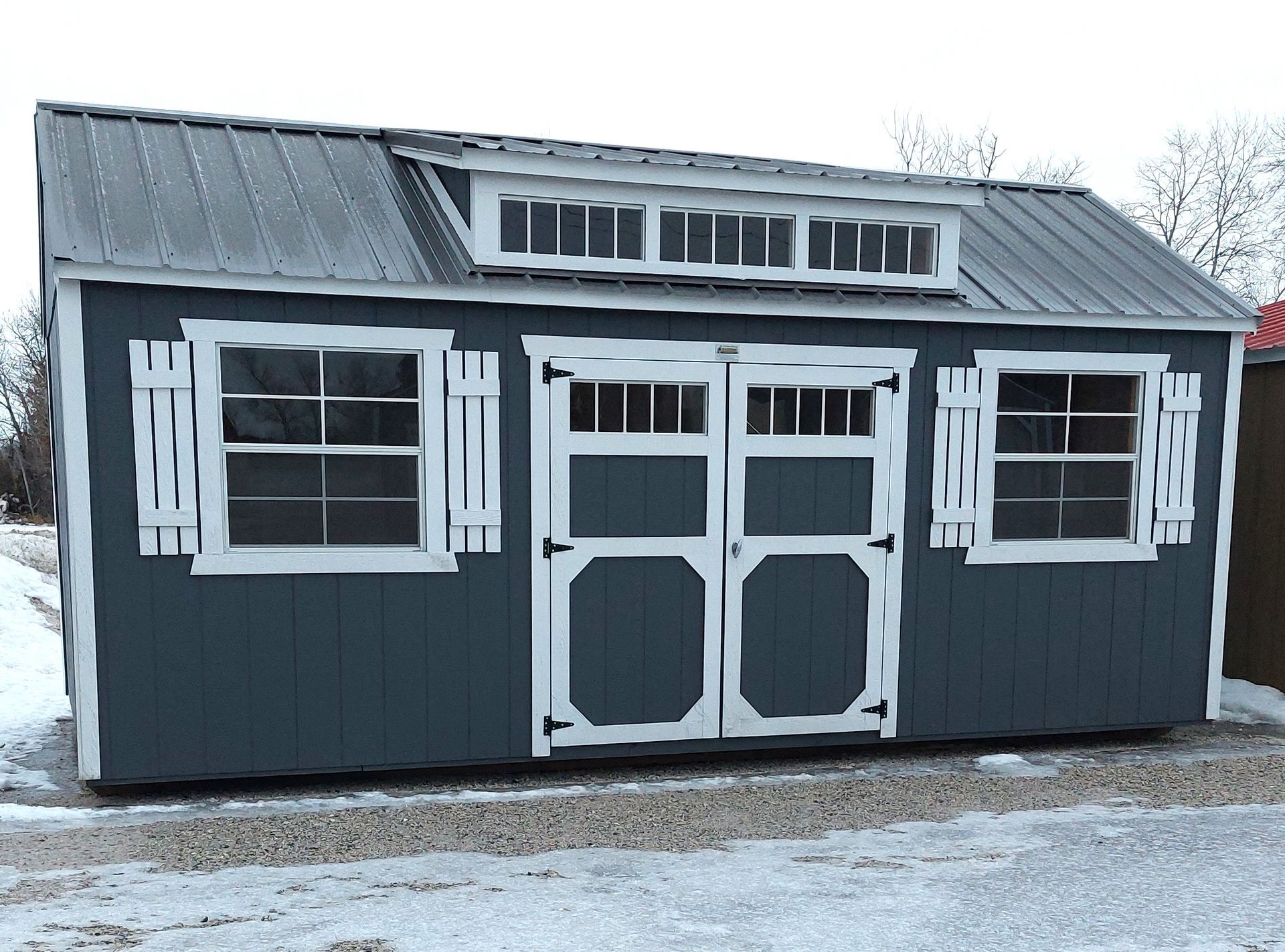 A dark blue wooden shed with white trim, a metal roof, two windows with shutters, and a central double door.