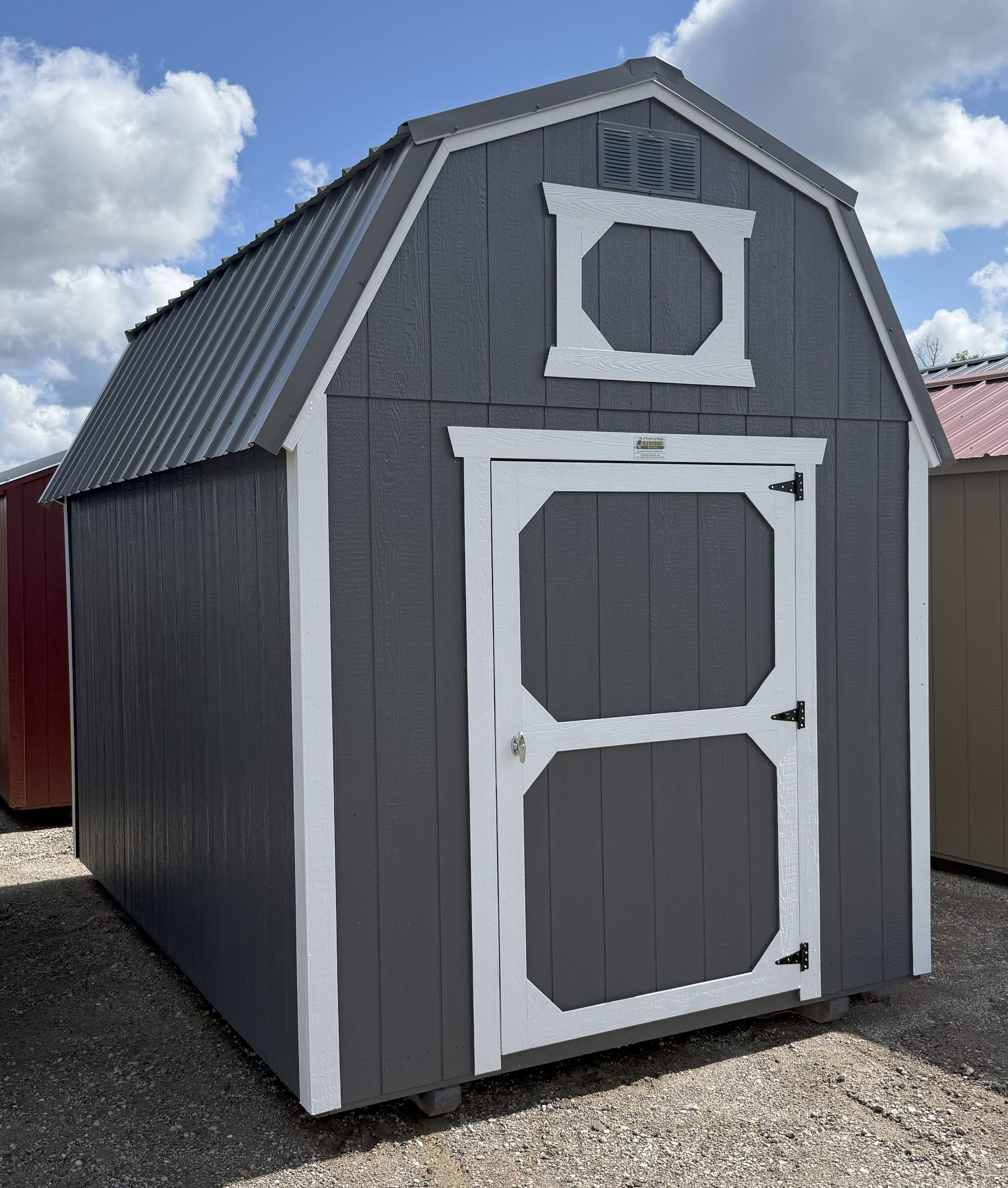 A gray, shed-style structure with a gambrel roof, white trim, a single door, and a small vent on a gravel lot.