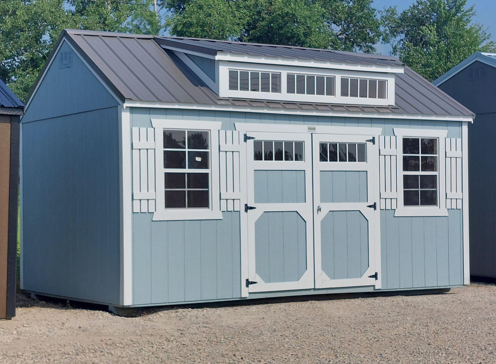 Light blue storage shed with white trim, a metal roof, transom windows, double doors, and shuttered windows on gravel.