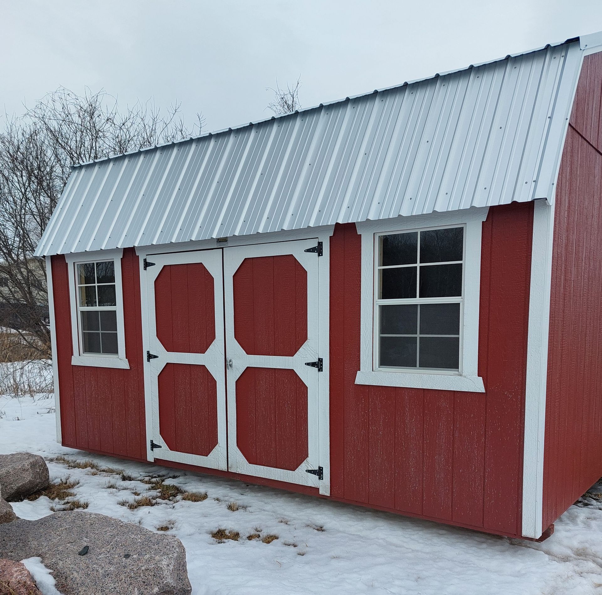 A red storage shed with white trim, double doors, and two windows, situated outdoors in a snowy landscape.