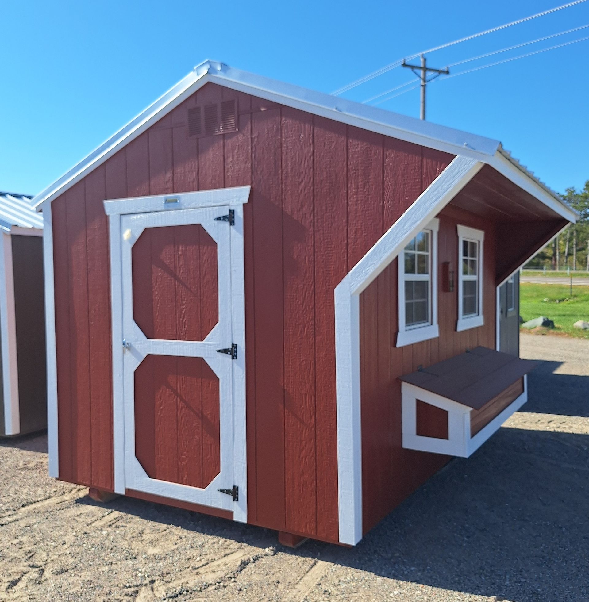 A red and white wooden shed with a side-mounted nesting box and windows, situated on a gravel lot under a blue sky.