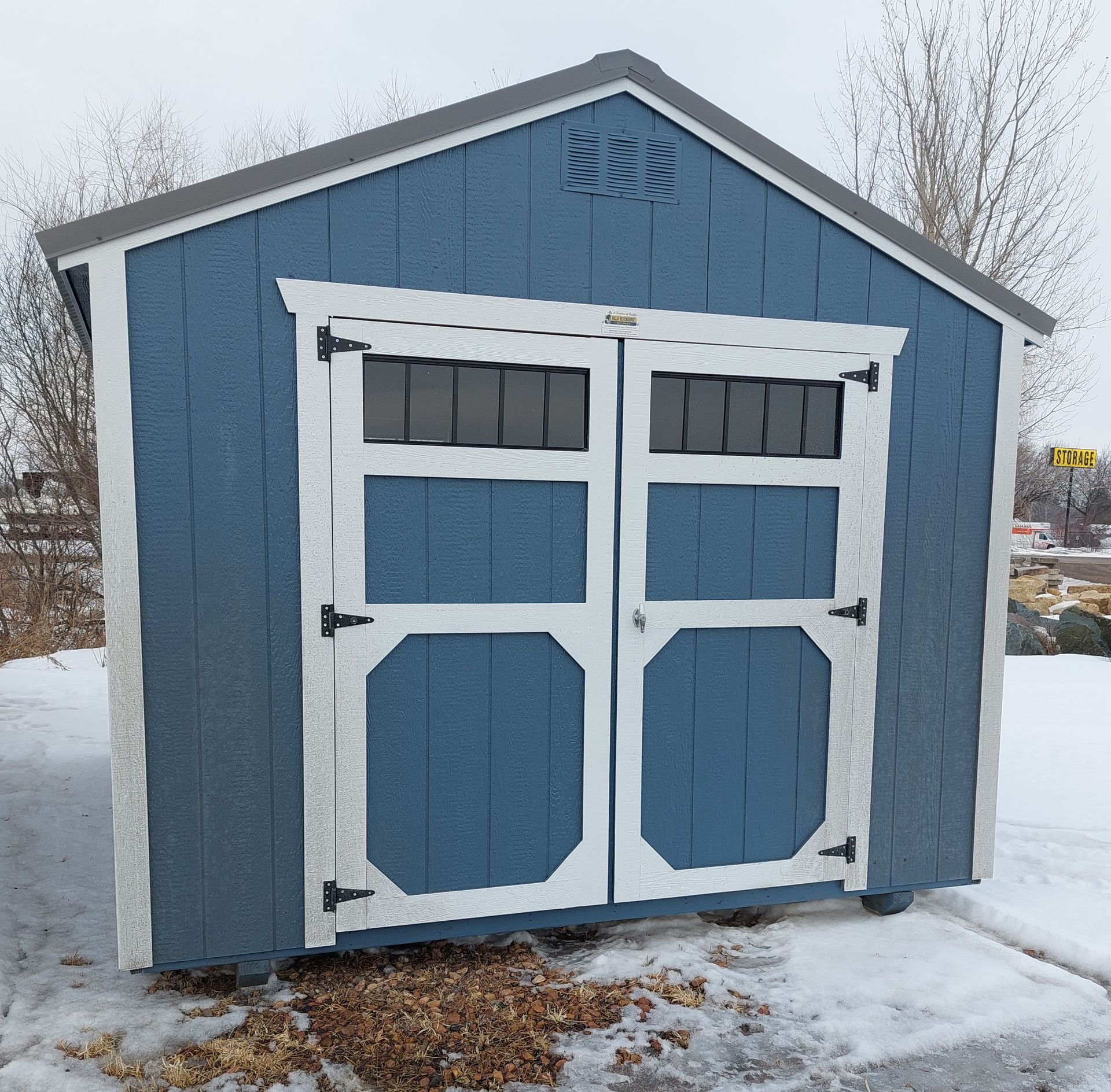 A blue and white wooden storage shed with double doors and transom windows, sitting on snowy ground.