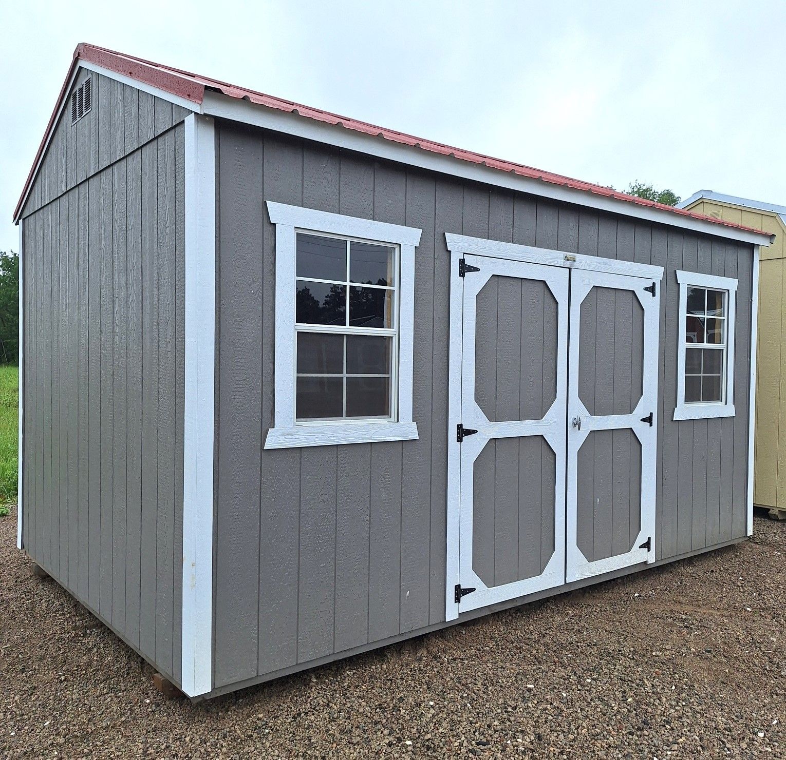 Gray shed with a red metal roof, white trim, double doors, and two windows, sitting on a gravel lot.