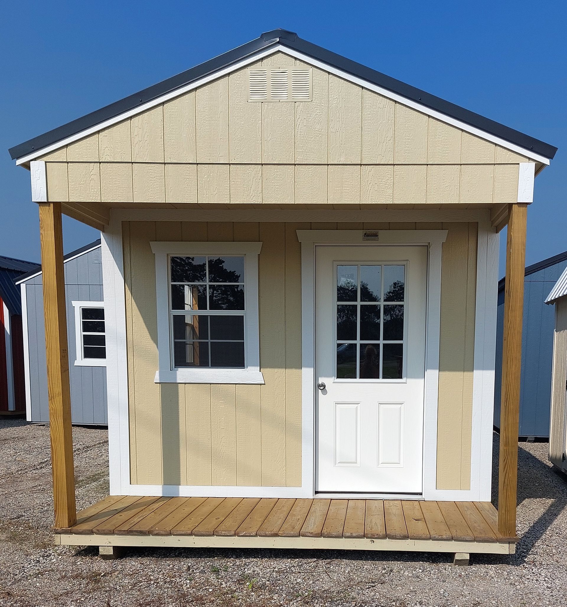 A tan, wooden shed with a small front porch, a single window, a white door with glass panels, and a gabled metal roof.