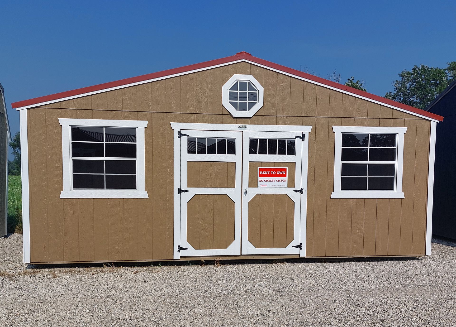A tan wooden shed with a red roof, white trim, two windows, double doors, and an octagonal window on a gravel lot.