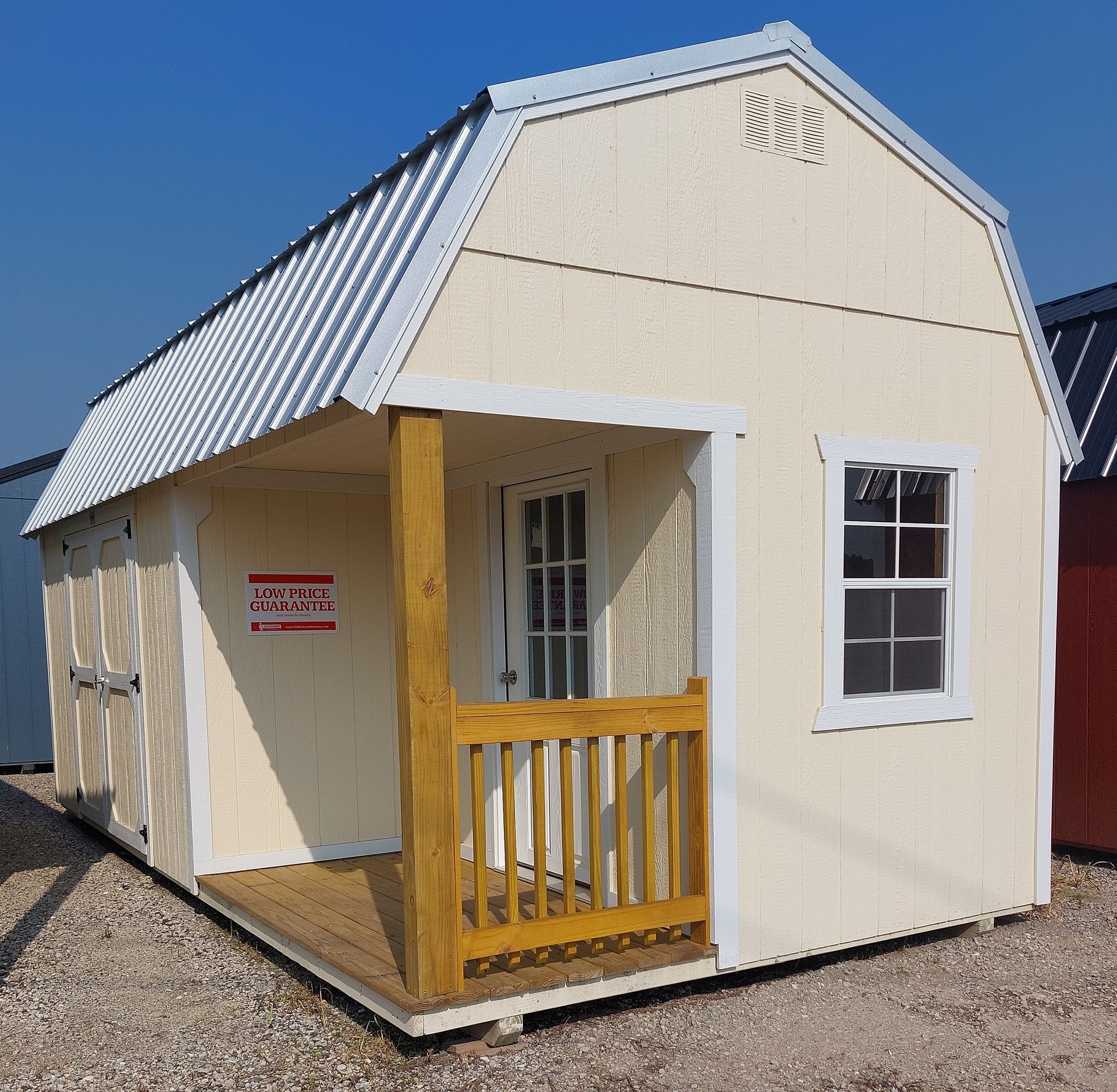 A yellow wooden shed with a metal gambrel roof, a front porch, a single door with windows, and a side-mounted barn door.