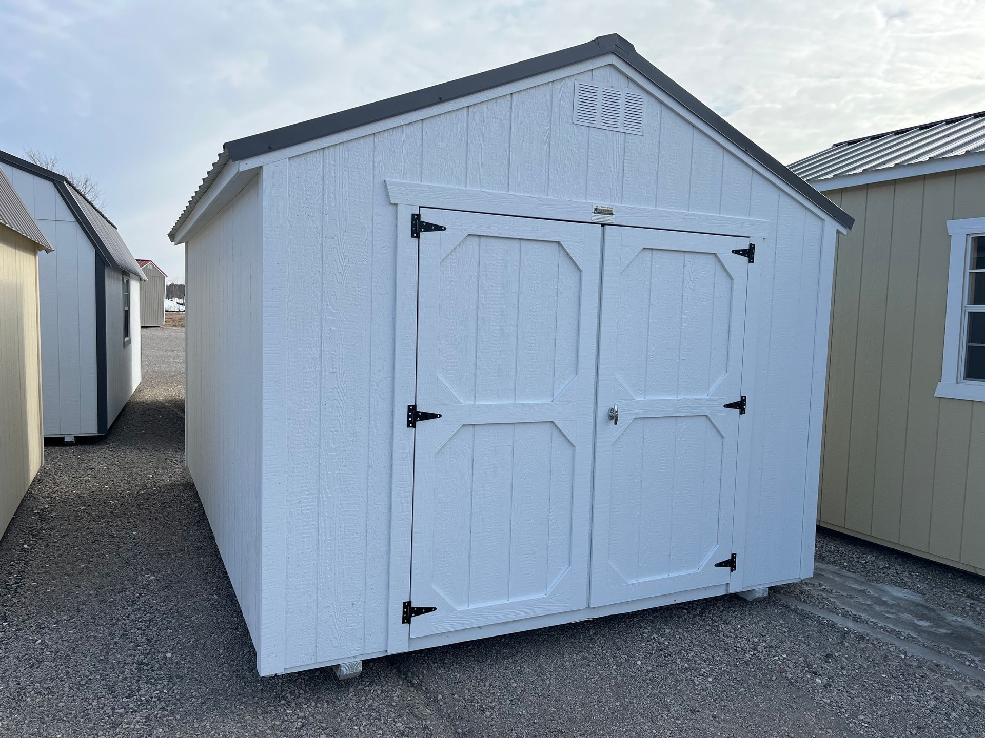A white wooden storage shed with double doors and a dark grey roof, positioned on a gravel lot next to other sheds.